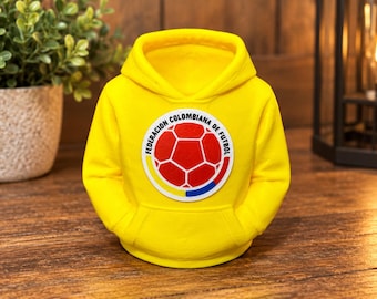 Puede incluir: Una sudadera con capucha en miniatura de color amarillo brillante que presenta un parche circular central con un gráfico de balón de fútbol rojo y el texto "FEDERACION COLOMBIANA DE FUTBOL" en negro, con bolsillo delantero.