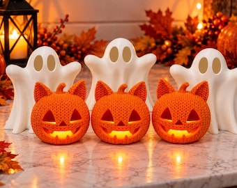Puede incluir: Tres calabazas de Halloween naranjas con orejas de gato e interiores brillantes están dispuestas en fila. Detrás hay tres figuras de fantasmas blancas. Las calabazas presentan ojos, narices y bocas dentadas talladas con textura.