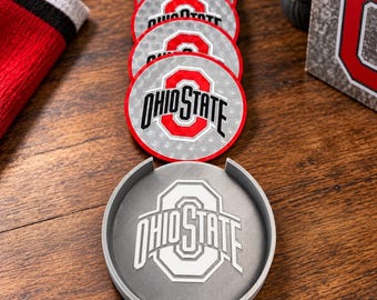 Puede incluir: Una pila de cuatro posavasos circulares con el logotipo rojo y negro de Ohio State sobre un fondo plateado, acompañada de un soporte circular gris con el logotipo blanco de Ohio State en relieve en la base.