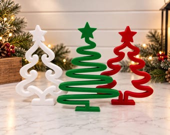 Puede incluir: Tres decoraciones de árbol de Navidad de pie en blanco, verde y rojo. Cada árbol presenta un diseño de línea continua estilizada con una estrella en la punta y una base rectangular plana, hecho de un material sólido con acabado mate.
