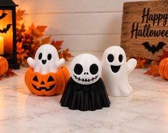 Puede incluir: Tres figuras con temática de Halloween: un fantasma blanco sentado dentro de una calabaza naranja, un personaje central con cabeza de calavera blanca y traje negro, y un fantasma blanco con una amplia sonrisa, sobre una superficie marmolada.