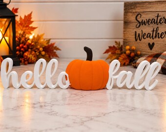 Puede incluir: Un letrero en letra cursiva blanca que dice hello fall con una calabaza de color naranja brillante y tallo marrón oscuro en el centro. El objeto decorativo está formado por letras individuales y una figura de calabaza.