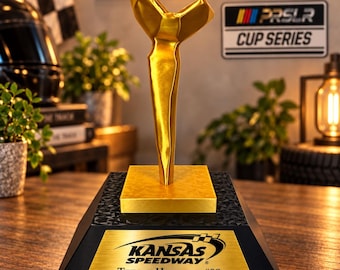 Puede incluir: Un trofeo de color dorado con una figura abstracta y angular montada sobre una base cuadrada. El pedestal negro en forma de pirámide muestra una placa dorada con el texto: KANSAS SPEEDWAY, TEEJAY HANDLEY #08, PIT ROAD SPEED LIMIT RACING, SEPTEMBER 9, 2026.