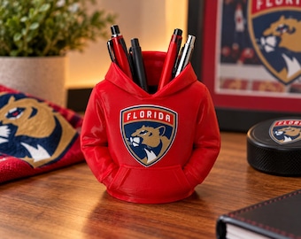 Könnte beinhalten: Ein roter Schreibtisch-Organizer in Form eines Miniatur-Kapuzenpullovers mit dem Logo des Eishockeyteams Florida Panthers auf der Brust. Der Organizer verfügt über eine Kapuze und eine Vordertasche und enthält derzeit vier Stifte.