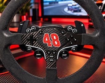Puede incluir: Un volante de carreras negro con una placa central de fibra de carbono que muestra el número 48 en rojo. El volante incluye varios botones etiquetados como WIP, N, FL, PL, BOX, P, CAM, RADIO, S1, S2, HOME, MENU, START y R, además de levas.