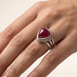 May include: A silver ring featuring a large, triangular, deep red cabochon gemstone set in a decorative bezel with beaded and twisted wire detailing. The band consists of three separate silver wires that merge into a single shank at the base.