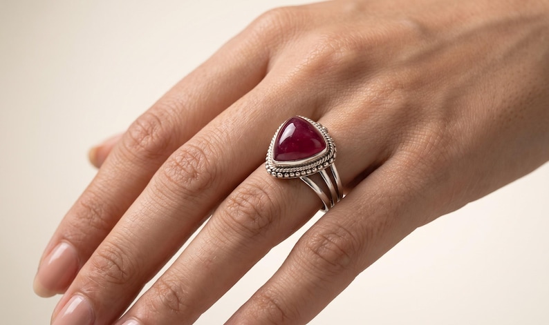 May include: A silver ring featuring a large, triangular, deep red cabochon gemstone set in a decorative bezel with beaded and twisted wire detailing. The band consists of three separate silver wires that merge into a single shank at the base.