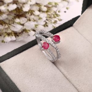 Puede incluir: Un anillo de plata con dos piedras preciosas rojas y pequeñas piedras transparentes. El anillo tiene un diseño de doble banda, con las piedras preciosas engastadas en la parte superior de cada banda. El anillo se muestra en una caja de joyería.