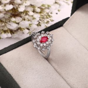 May include: A silver ring with a central oval ruby, encircled by small, clear gemstones. The ring has a floral or starburst design. It's presented in a jewellery box with a neutral-toned interior. The ring's design is intricate.