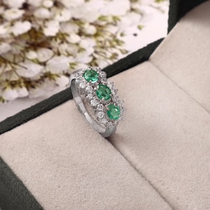 Può includere: Un anello color argento con tre pietre preziose verdi a taglio ovale disposte in fila, circondate da un alone di piccole pietre bianche rotonde e trasparenti incastonate nella fascia metallica, creando un design a grappolo sulla parte superiore.