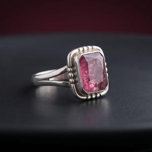 Op de afbeelding: Een zilveren ring met een rechthoekige, gefacetteerde roze edelsteen in een zetting. De metalen zetting is voorzien van decoratieve kraaldetails langs de randen van de steen en de ringband heeft een gespleten ontwerp.