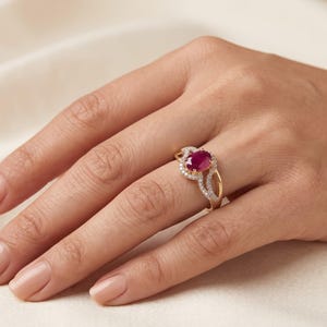 May include: A gold ring featuring a central oval-cut deep red ruby gemstone. The ruby is surrounded by a halo of small, clear, round-cut white stones. The band is crafted from polished yellow gold and has a split shank design with stone accents.