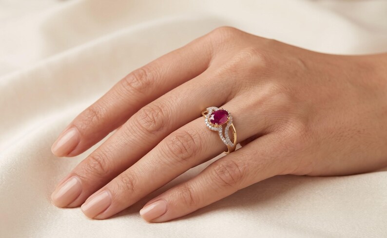 May include: A gold ring featuring a central oval-cut deep red ruby gemstone. The ruby is surrounded by a halo of small, clear, round-cut white stones. The band is crafted from polished yellow gold and has a split shank design with stone accents.