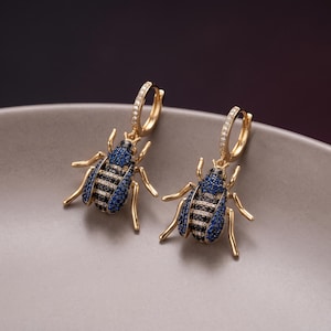 Könnte beinhalten: Ein Paar goldene Creolen mit baumelnden käferförmigen Anhängern. Die Körper der Käfer sind mit kleinen blauen und schwarzen Steinen besetzt, während die goldenen Creolen auf der Vorderseite mit winzigen klaren Steinen verziert sind.