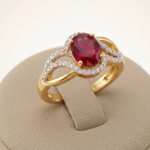 May include: A gold ring featuring a central oval-cut deep red ruby gemstone surrounded by a halo of small white diamonds. The band has a split-shank design with additional small white diamonds set along the curving gold arms of the ring setting.