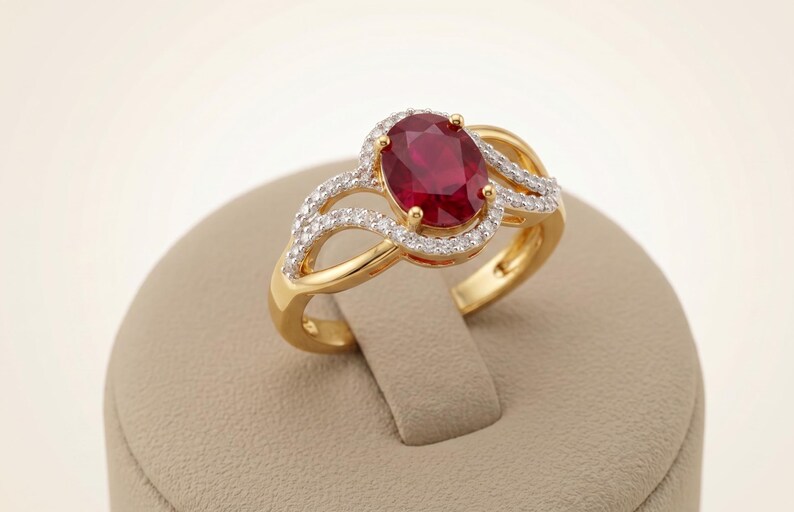 May include: A gold ring featuring a central oval-cut deep red ruby gemstone surrounded by a halo of small white diamonds. The band has a split-shank design with additional small white diamonds set along the curving gold arms of the ring setting.