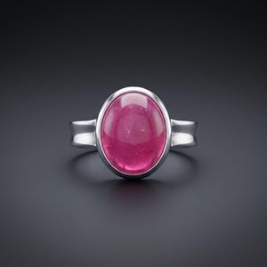 May include: A silver ring featuring a large, oval-shaped, cabochon-cut pink gemstone set in a smooth, polished metal bezel. The band is solid silver with a simple, clean design that tapers slightly as it connects to the central stone setting.