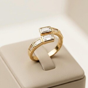 Puede incluir: Un anillo de oro tipo bypass con una serie de piedras transparentes de corte baguette rectangular a lo largo de la banda. El diseño incluye dos extremos superpuestos, cada uno terminado con una piedra, creando un aspecto estructurado.