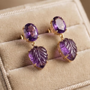 Könnte beinhalten: Ein Paar Ohrhänger mit oval geschliffenen, violetten Amethyststeinen an der Oberseite, die mit geschnitzten Amethystblättern an der Unterseite verbunden sind. Die Steine sind in goldfarbenes Metall gefasst und bilden ein hängendes Design.