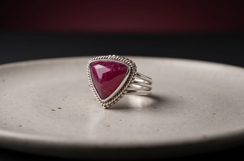 May include: A silver ring featuring a large, triangular, faceted deep pink gemstone set in a bezel with a twisted rope-style border. The band consists of three separate silver wires that join together at the base of the setting.