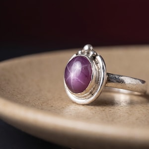 May include: A silver ring featuring a round, cabochon-cut purple star sapphire gemstone. The stone displays a distinct six-rayed star pattern on its surface. The metal setting is smooth and polished, with a small decorative bead detail at the top.