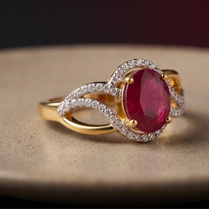 May include: A gold ring featuring a large, oval-cut deep red gemstone at the center, surrounded by a halo of small, clear white stones. The band splits into two delicate, curved arms that are also encrusted with small, clear white stones.