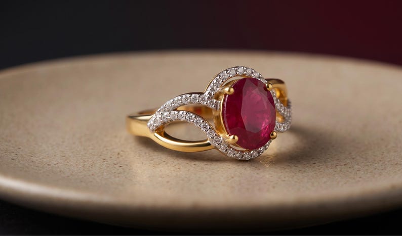May include: A gold ring featuring a large, oval-cut deep red gemstone at the center, surrounded by a halo of small, clear white stones. The band splits into two delicate, curved arms that are also encrusted with small, clear white stones.