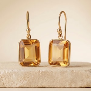 May include: A pair of gold drop earrings featuring rectangular, emerald-cut yellow citrine gemstones set in polished gold frames. The earrings hang from simple gold shepherd hook wires, showcasing the translucent, warm amber hue of the stones.