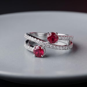 May include: A silver bypass ring featuring two round-cut red rubies set at the ends of the crossover band. The band is encrusted with small, clear pavé-set stones that extend along the split shank, creating a sparkling, layered appearance.