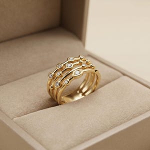 Op de afbeelding: Een gouden ring met meerdere banden, bestaande uit verschillende dunne, met elkaar verbonden ringen versierd met kleine, ronde, heldere stenen in een zetting. De ring wordt gepresenteerd in een open juwelendoos in neutrale tinten.