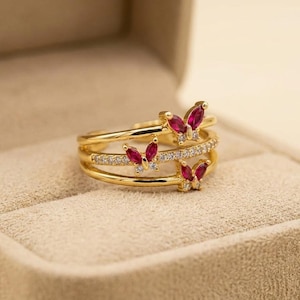 May include: A gold-toned ring featuring three butterfly motifs with marquise-cut red stones and small clear accent stones. The band consists of three parallel gold-toned wires, with the butterflies positioned along the top and middle sections.