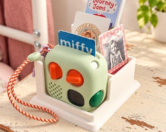 May include: A light green portable music player with orange knobs and a speaker grille, sitting in a white plastic organizer stand. The stand holds several rectangular cards, including ones labeled Journey Jams, Little Pilgrim, and miffy.
