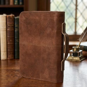 May include: A brown leather case with a handle, likely for documents or a tablet. The case has a stitched horizontal line across the middle and a textured surface. The handle is attached to the side with a leather strap.