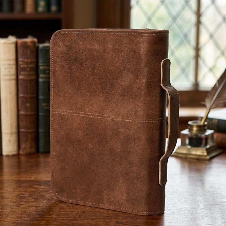 May include: A brown leather case with a handle, likely for documents or a tablet. The case has a stitched horizontal line across the middle and a textured surface. The handle is attached to the side with a leather strap.
