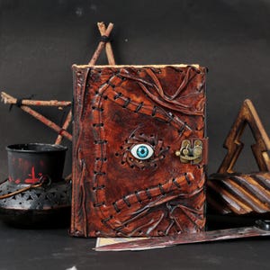 May include: A brown leather journal with a stitched design and a blue eye in the center. The journal has a clasp closure and is surrounded by other decorative items, including a small cauldron and a wooden structure.