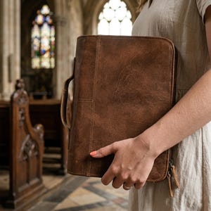 May include: A brown leather portfolio with a handle and zipper closure. The portfolio has a stitched design and is held in front of a church interior with stained glass windows.