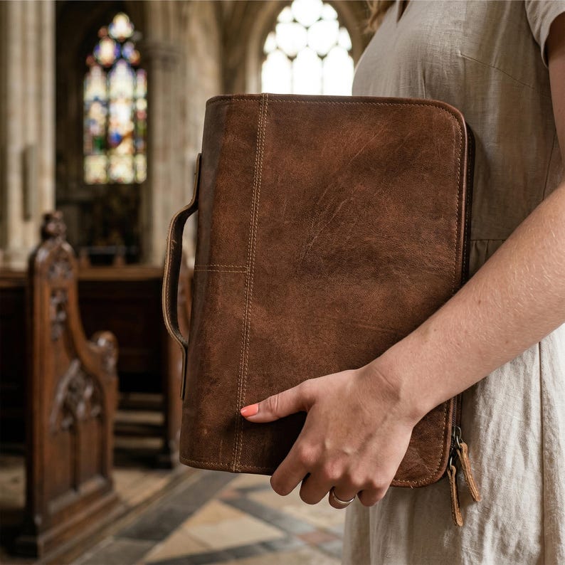 May include: A brown leather portfolio with a handle and zipper closure. The portfolio has a stitched design and is held in front of a church interior with stained glass windows.