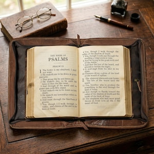 May include: An open Bible, displaying Psalm 23, rests in a brown leather case on a wooden surface. A pair of round eyeglasses and a closed book are visible in the background, along with a pen and inkwell. The scene evokes a sense of study and reflection.