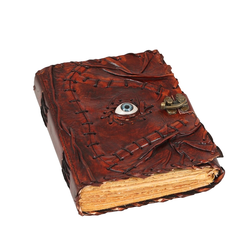 May include: A brown leather-bound book with a stitched design and a brass clasp. The cover features a blue and white eyeball and the pages are aged. A unique, handcrafted item.