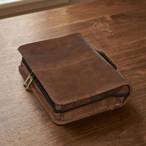 May include: A brown leather Bible cover with a zipper closure and a handle. The cover is rectangular and has a stitched design. The leather has a distressed look, and the zipper is gold-colored. The cover is sitting on a wooden surface.