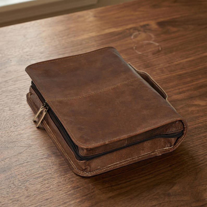 May include: A brown leather Bible cover with a zipper closure and a handle. The cover is rectangular and has a stitched design. The leather has a distressed look, and the zipper is gold-colored. The cover is sitting on a wooden surface.