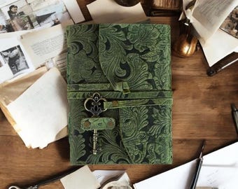 May include: A rectangular green journal featuring an embossed floral pattern across the cover. A leather strap wraps around the center, secured by a decorative metal key-shaped clasp. The journal is positioned vertically with a textured surface.