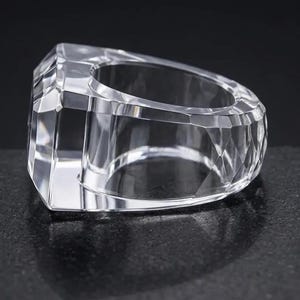 Può includere: Un anello trasparente e incolore caratterizzato da un design geometrico sfaccettato con molteplici superfici angolari che catturano e riflettono la luce. La fascia è spessa e larga, con un aspetto lucido simile al vetro e bordi netti.