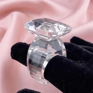 May include: A large, clear, faceted ring made of transparent material. The ring features a prominent, rectangular-cut stone setting atop a wide, multi-faceted band. The design is entirely colourless and reflective, showcasing sharp geometric edges.