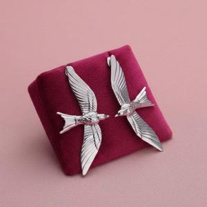 Op de afbeelding: Een paar oorbellen van zilverkleurig metaal in de vorm van vliegende zwaluwen met gedetailleerd gegraveerde vleugelveren. De oorbellen zijn geplaatst op een klein, vierkant presentatiekussentje van diepmagenta fluweel met een zachte textuur.