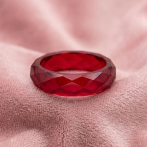 Könnte beinhalten: Ein facettierter, tiefroter Ring. Der Ring hat ein geometrisches Design mit mehreren flachen Oberflächen, wodurch ein funkelnder Effekt entsteht. Der Ring liegt auf einem weichen, rosa Hintergrund.