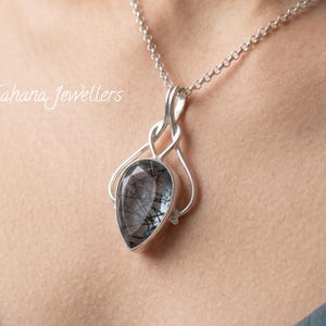Könnte beinhalten: Eine silberne Halskette mit Anhänger, die einen tropfenförmigen Edelstein mit schwarzen Einschlüssen zeigt. Der Anhänger hängt an einer feinen Kette. Der Text "Sahana Jewellers" ist sichtbar.