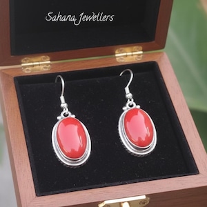 May include: A pair of red oval earrings with a silver-colored border, displayed in a wooden jewelry box with the text "Sahana Jewellers". The earrings have a hook closure and are set against a black velvet interior.