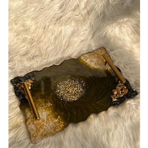 Może przedstawiać: Dekoracyjny podkład z unikalnym wzorem. Taca ma ciemnozieloną i złotą powierzchnię z żywicy ze złotymi uchwytami. Krawędzie zdobią złote akcenty i czarne kamienie, tworząc elegancki i artystyczny wygląd.