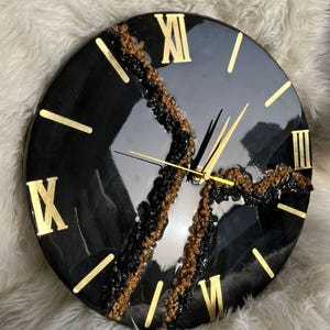 Può includere: Un orologio da parete rotondo nero con numeri romani e lancette dorate. Il quadrante presenta un design decorativo e strutturato nei toni del nero, marrone e oro. È un oggetto di arredamento.